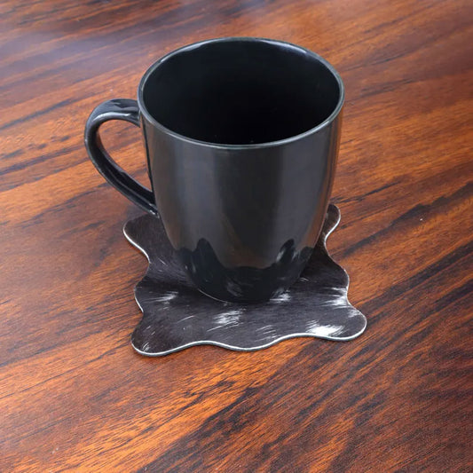 Hair-on leather coasters placed on a wooden bar counter beneath glasses of whiskey and cocktails, complementing rustic bar décor and evoking a warm, inviting setting.

